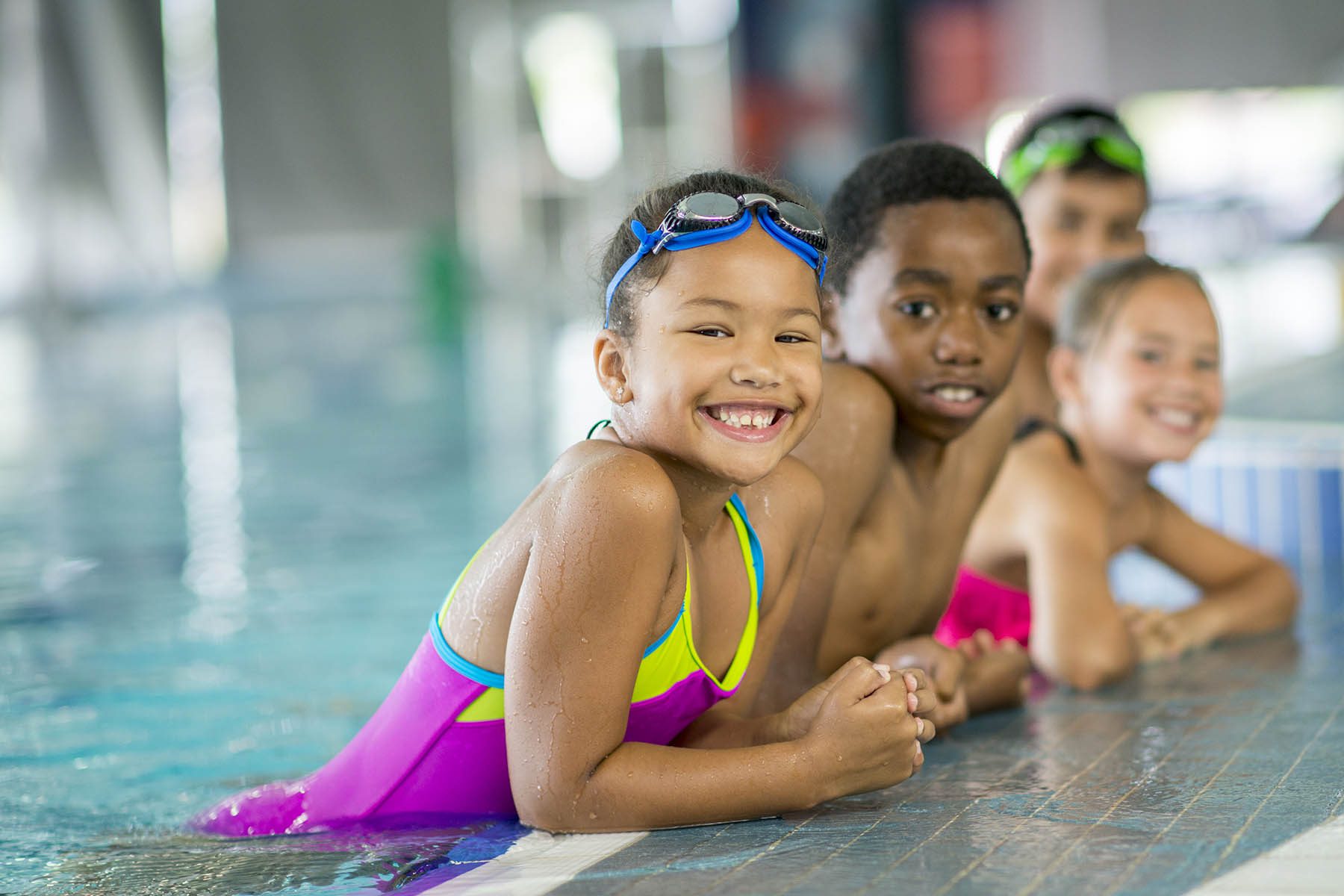 A multi-ethnic group of elementary children are taking swimming lessons together at the pool. They are smiling and looking at the camera.