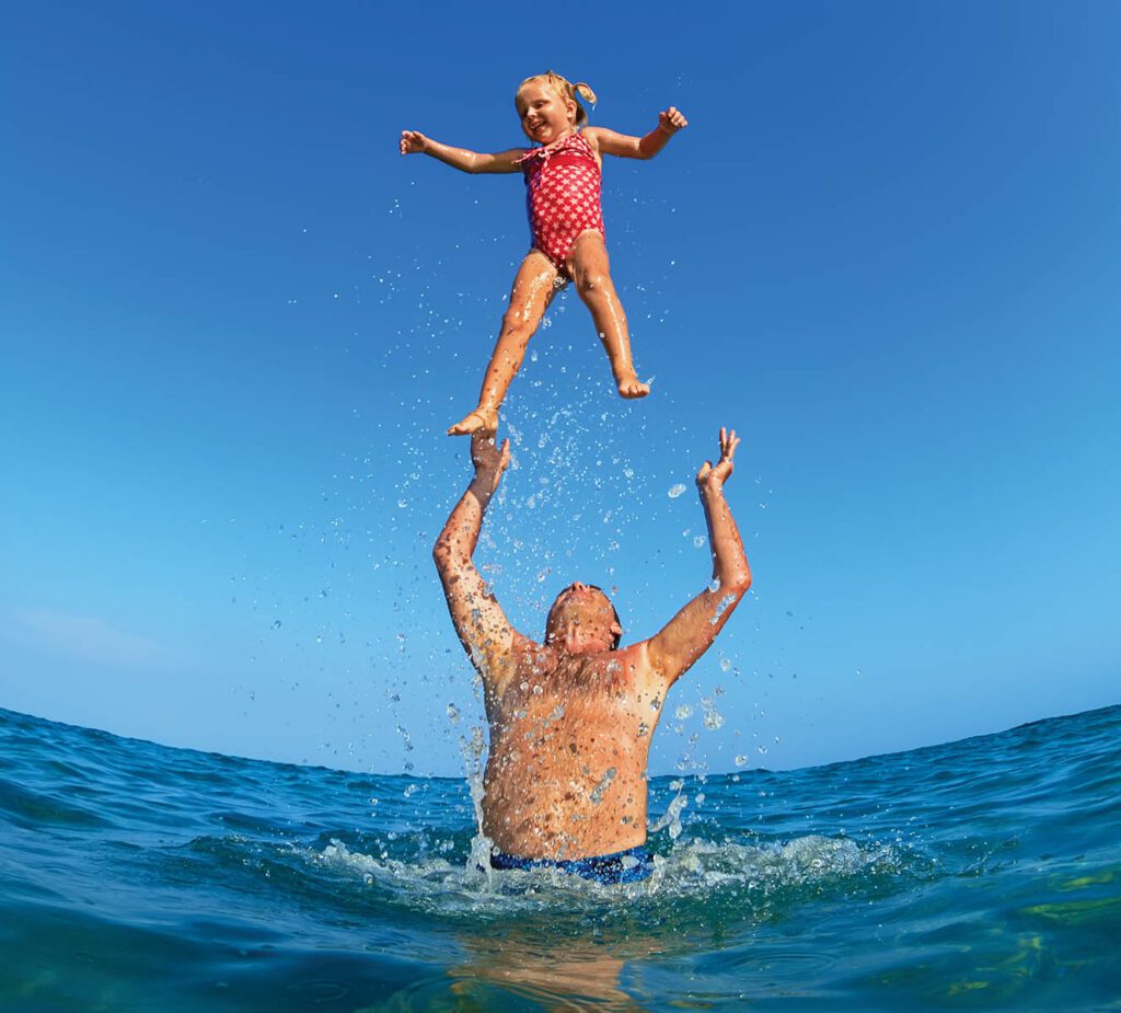 Grandfather tossing up the high air with water splashes joyful baby girl on sea beach. Outdoors child activity, active healthy lifestyle, swimming with fun on summer family vacation on tropical island.